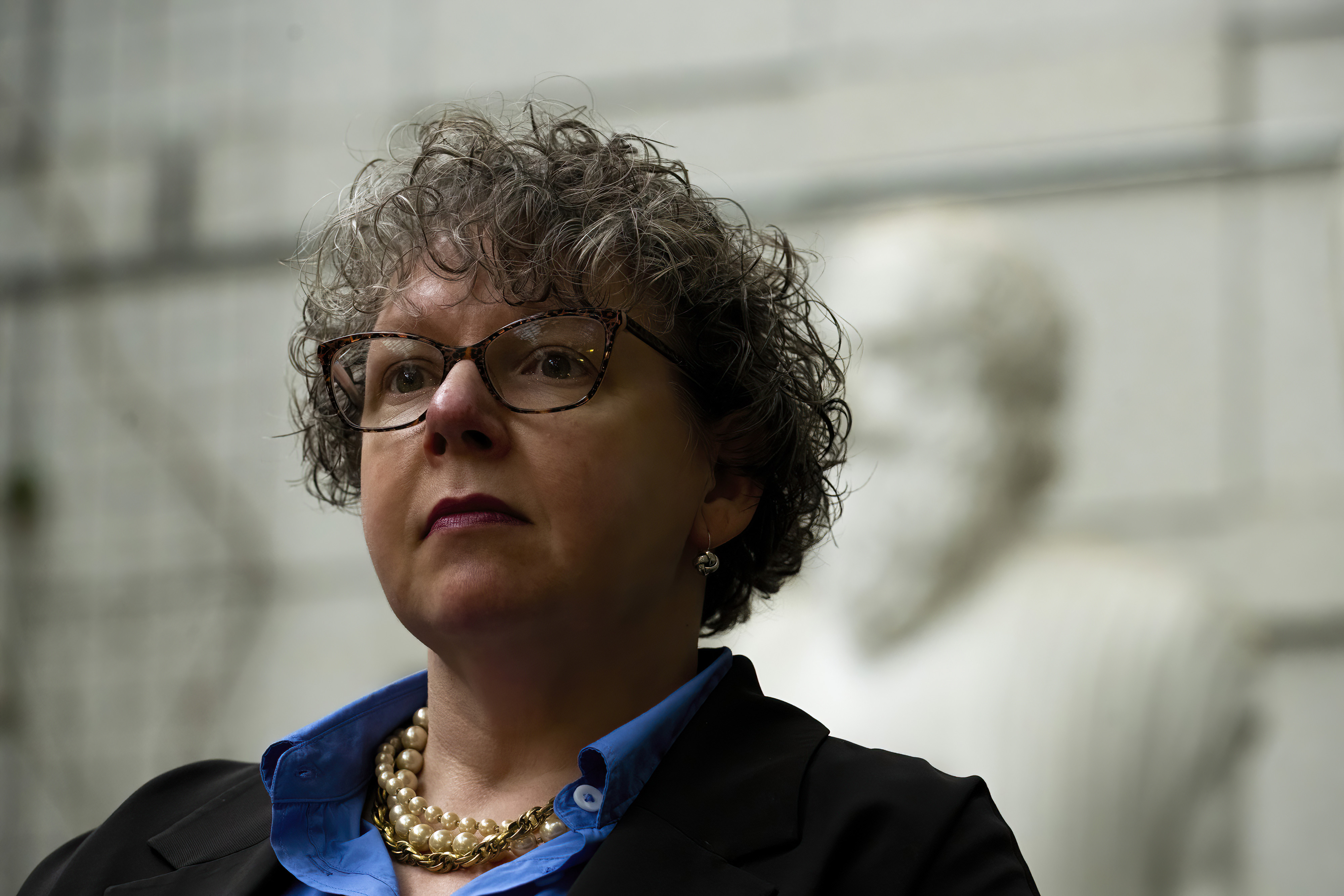 Sarah Elaine Eaton - Short curly hair, glasses, wearing a necklace, a blue shirt, and a black jacket. The white and background is blurred.