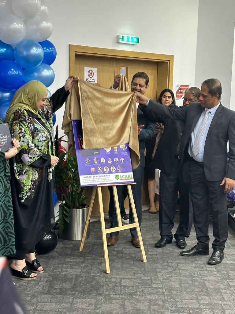 Unveiling of the Handbook of Academic Integrity (2nd ed.) Two people are removing a gold-coloured cover from a poster featuring the book cover. People are standing in the background. There are blue and white balloons on the left edge of the frame.